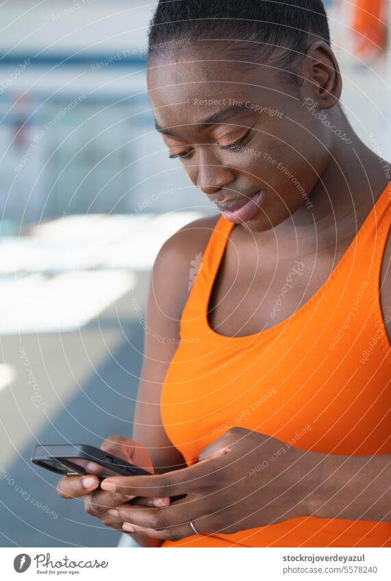 Schwarze junge Frau, die ihr Smartphone benutzt und auf einer Bank am Schwimmbad sitzt Frauen Person Lifestyle Telefon Mobile schwarz erhitzt Badeanzug orange