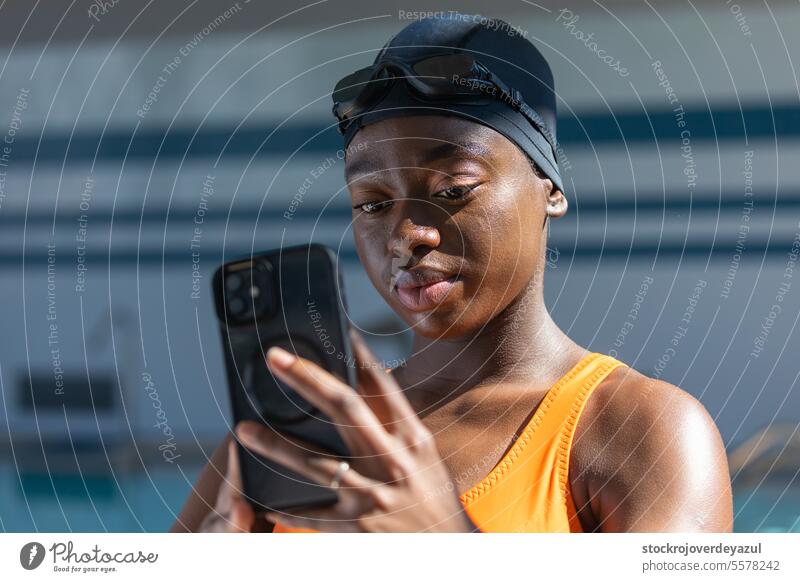 Schwarze junge Frau hält ihr Smartphone in der Hand, während sie am Swimmingpool steht schwarz Schwimmbad erhitzt Badeanzug orange Gesundheit Lächeln