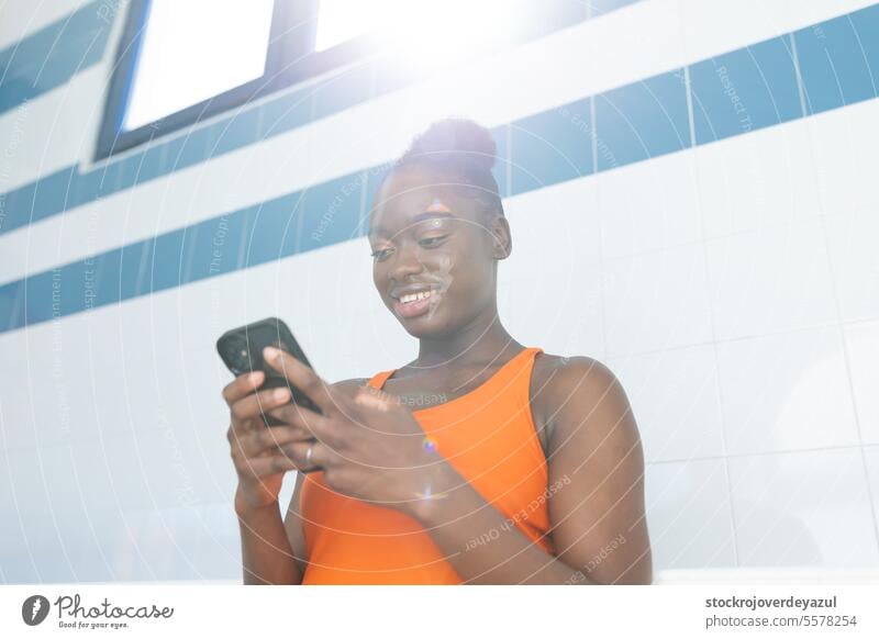 Schwarze junge Frau, die ihr Smartphone unter einem schönen Licht am Schwimmbad benutzt schwarz erhitzt Badeanzug orange Gesundheit blau Lifestyle Sport