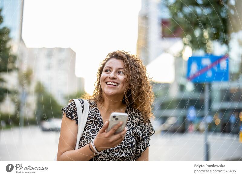 Porträt einer lächelnden jungen Frau, die in der Stadt ein Mobiltelefon benutzt junger Erwachsener Straße außerhalb Selbstvertrauen Lächeln genießen Lachen