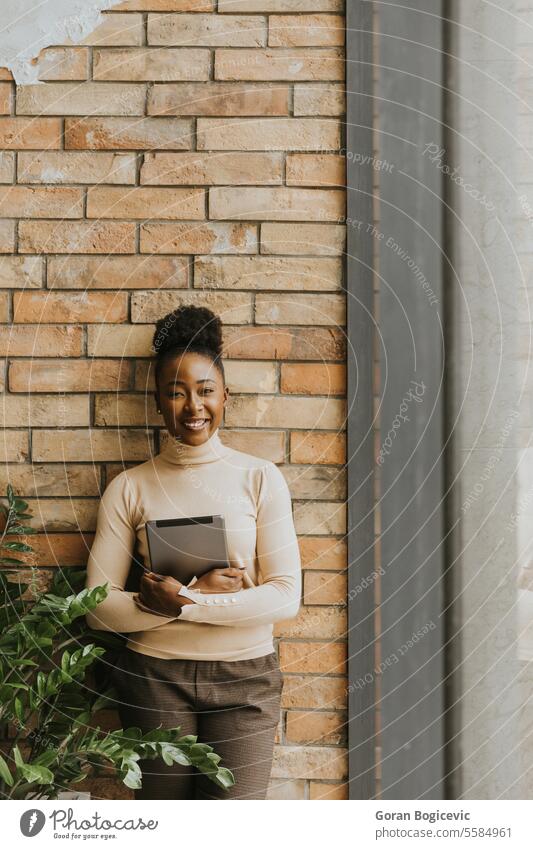 Eine junge afroamerikanische Geschäftsfrau mit digitalem Tablet steht an der Backsteinmauer in einem Büro im Industriestil. Erwachsener Afrikanisch Amerikaner