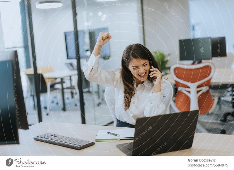 Junge Geschäftsfrau, die ein Mobiltelefon benutzt, während sie an einem Laptop in einem modernen Büro arbeitet Beruf Smartphone Frau jung Glück sprechend