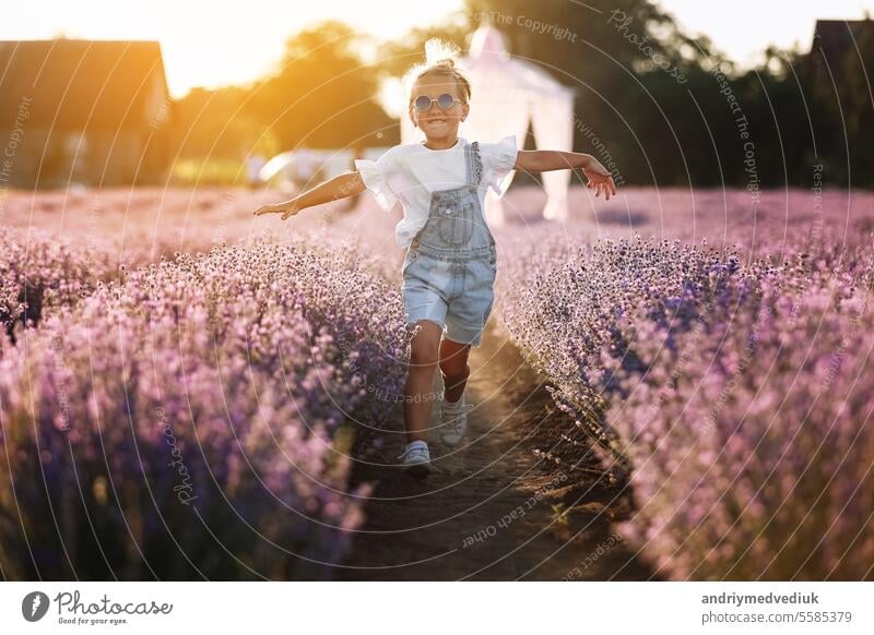 glückliches Kind Mädchen ist in Lavendelfeld auf Sonnenuntergang zu Fuß. Lächelnde Kind in Zuckerbrille, Jeans Overall hat Spaß in der Natur am Sommertag. Fröhlich kleines Mädchen. glückliche Kindheit Konzept