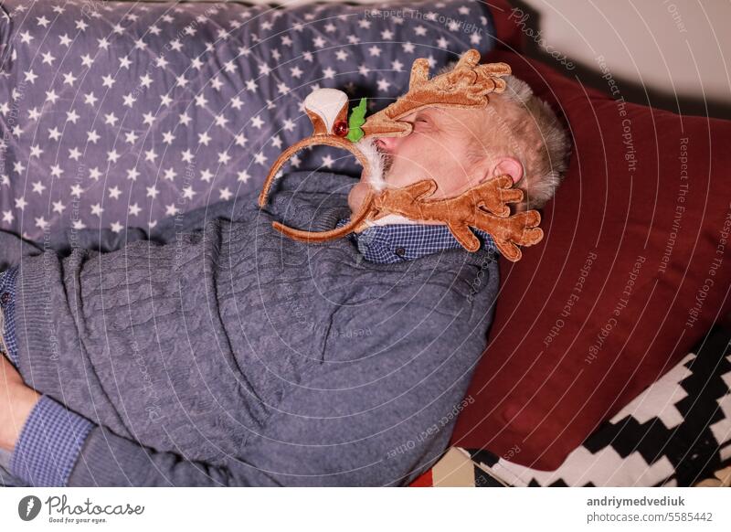 Mann von 50-60 Jahren, der auf einem Sofa schläft und eine Dekoration in Form von Hirschhörnern auf dem Kopf trägt. Mann ruht sich nach der Weihnachts- und Silvesterfeier aus