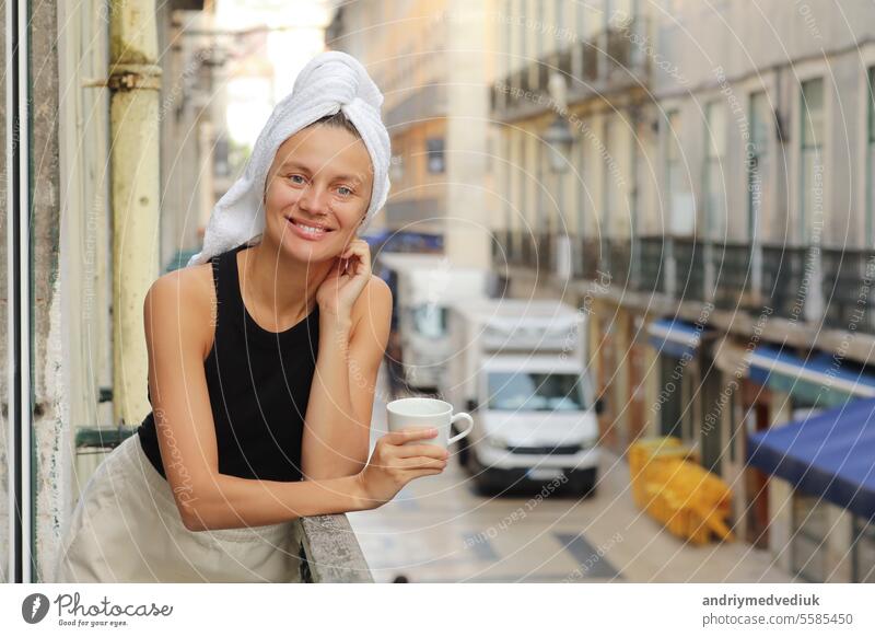 Junge schöne Frau in einem Handtuch auf dem Kopf trinkt Kaffee oder Tee und lächelnd niedlich stehend auf Balkon auf ueropean Straße Hintergrund. lifestyle Morgen. Konzept der inländischen oder Reisen Lebensstil.