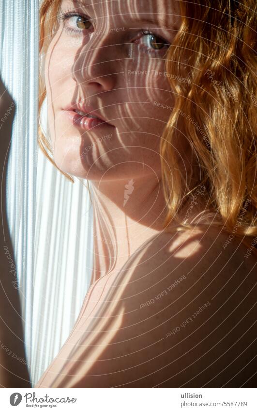 Attraktives sexy Porträt einer jungen schönen rothaarigen Frau, mit linearen Schattenstreifen auf dem Gesicht durch die weißen Fäden eines Fadenvorhangs