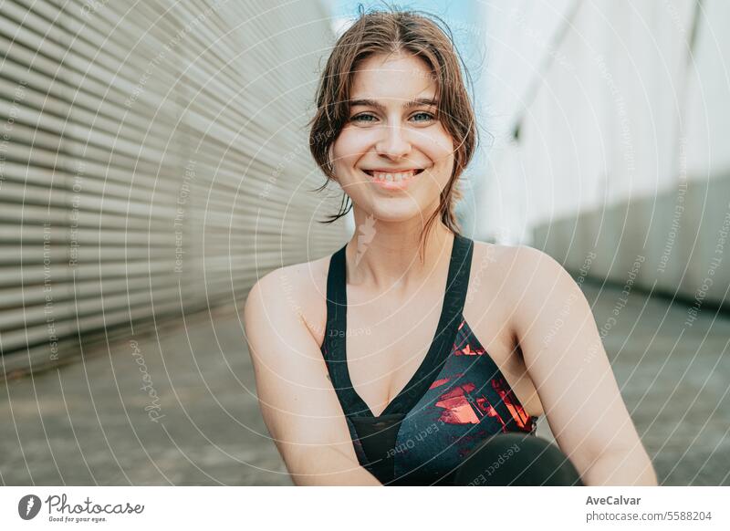 Eine fitte, lächelnde und fröhliche junge Frau in Sportkleidung sitzt auf dem Boden in einer städtischen Straßenszene. Schönheit Person Glück Porträt im Freien