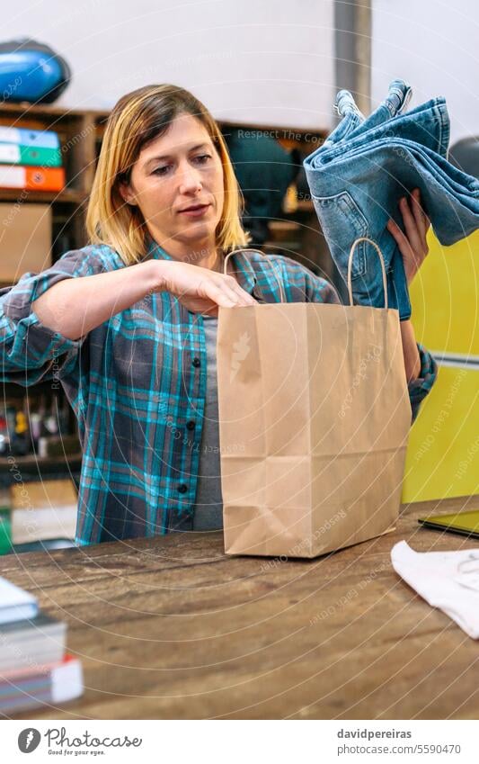 Konzentrierte Verkäuferin, die Jeans in eine ökologische Papiertüte für einen Online-Shop-Kunden steckt Konzentration Frau Werkstatt Assistent Setzen Jeanshose