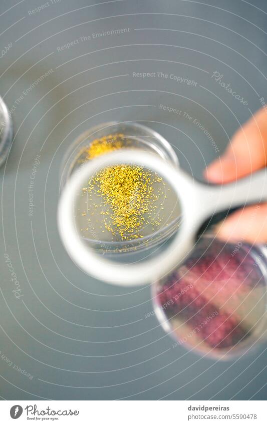 Eine Chemikerin hält eine Lupe in der Hand, um eine goldene Glitzerprobe auf einer Petrischale im Labor zu analysieren Frau Beteiligung vergrößernd Glas Analyse