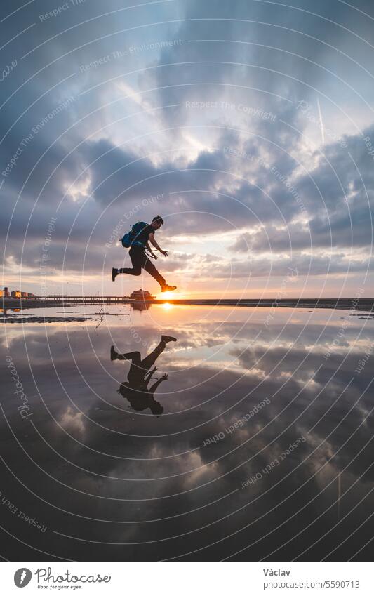 Braunhaariger Mann mit Rucksack, 25-29 Jahre alt, springt bei Sonnenuntergang in ein Wasserbecken am Strand. Er fängt die Sonne zwischen dem Boden und seinen Füßen ein. Oostende, Belgien