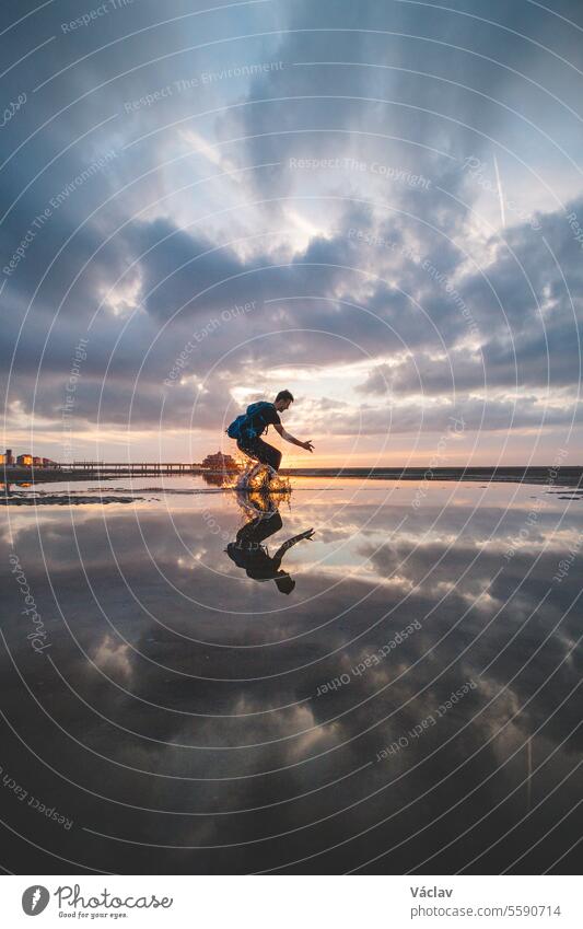 Braunhaariger Mann mit Rucksack, 25-29 Jahre alt, springt bei Sonnenuntergang in ein Wasserbecken am Strand. Er fängt die Sonne zwischen dem Boden und seinen Füßen ein. Oostende, Belgien