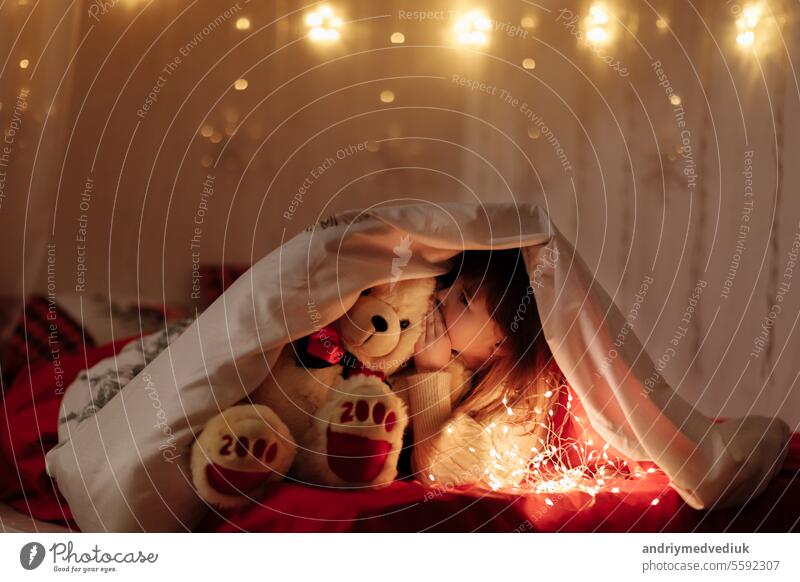 kleine niedliche lächelnde Baby-Mädchen mit weißen Teddybär späht aus unter einer Decke auf einem Bett zu Hause mit Beleuchtung Girlanden bei Dunkelheit. Kind schaut in die Kamera. Fairytale Kindheit mit Wunder Konzept