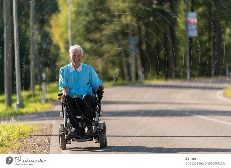 Lächelnde behinderte alte Frau in einem Rollstuhl auf der Landstraße Senior ungültig deaktiviert Person Autobahn Asphalt Straße fahren Freiheit älter Glück