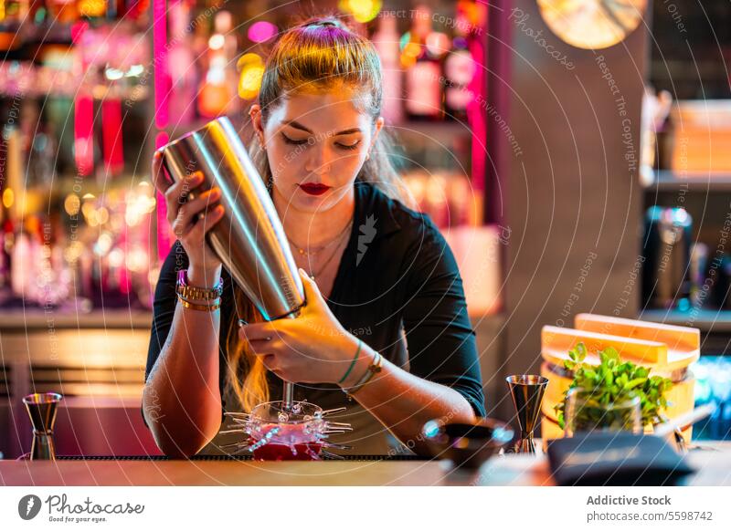 Fokussierte junge Bardame mit blondem Haar steht am Tresen und macht einen Cocktail an einer beleuchteten Bar Frau Barkeeperin Gießen Flasche Glas Alkohol