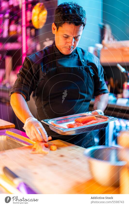 Fokussierter Koch in schwarzer Schürze und Handschuhen bei der Zubereitung eines orientalischen Abendessens mit Lachsfleisch am Tresen einer Sushi-Bar Mann