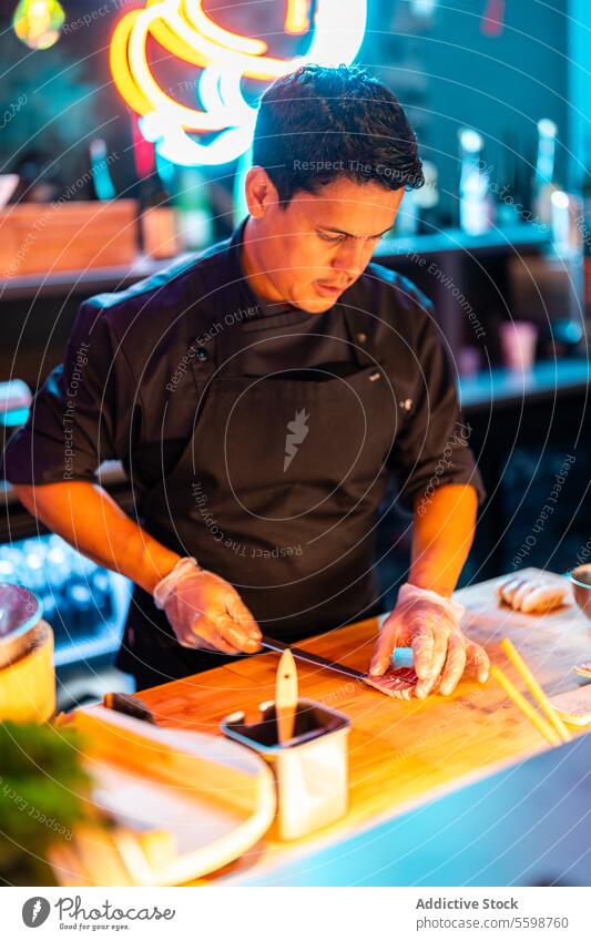 Fokus Männlicher Koch in Handschuhen und schwarzer Schürze steht beim Schneiden von Fleisch mit einem Messer auf dem Tresen einer modernen Sushi-Bar Mann