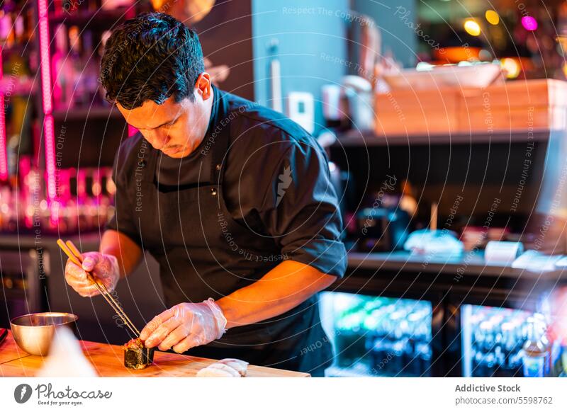 Fokussierte Köchin in schwarzer Schürze und Handschuhen bei der Arbeit an der Sushi-Bar mit Stäbchen für die Zubereitung frischer Sushi-Rollen Mann Küchenchef