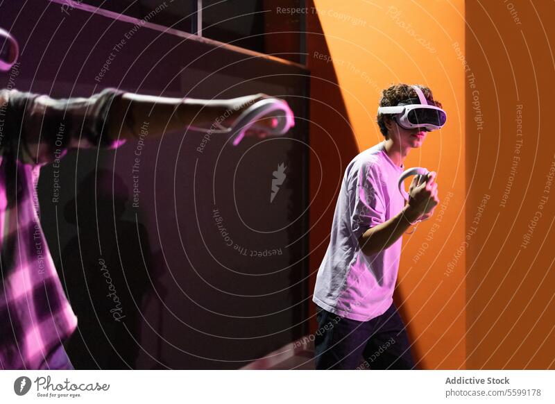 Seitenansicht von zwei Freunden, die in einem Spielbereich stehen und vollständig in eine Welt der virtuellen Realität eintauchen, VR-Headsets tragen und Bewegungssteuerungen in einem Raum mit Neonlicht halten
