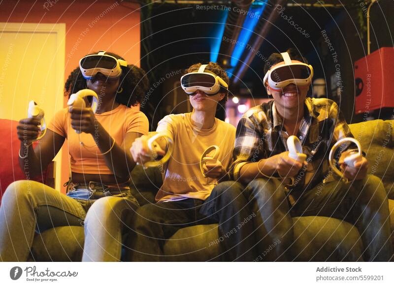Multiethnischer Freund, der in ein Virtual-Reality-Spiel vertieft ist, VR-Headsets trägt und Controller in der Hand hält, auf einer Couch in einem schwach beleuchteten Raum sitzt