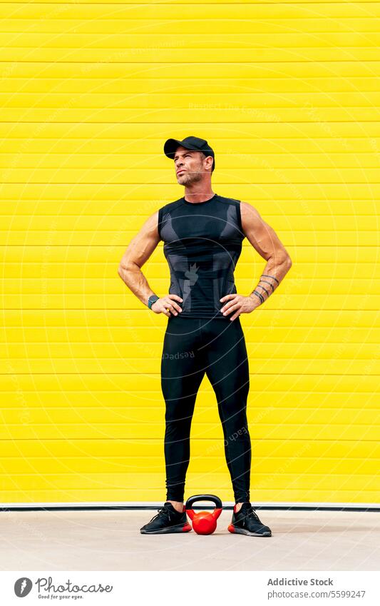 Ganzer Körper eines Sportlers in der Stadt während einer Pause Athlet Kettlebell Arme ausbreiten ruhen muskulös Mann Stehen Straße gelb Wand Training