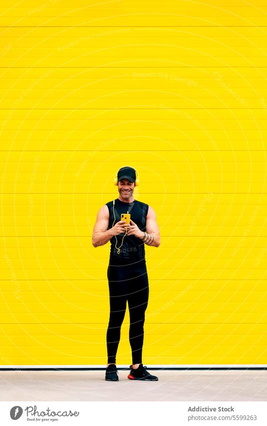 Selbstbewusster männlicher Sportler, der während des Trainings telefoniert Athlet Smartphone Lächeln zuhören Musik Kopfhörer Straße gelb Wand Ganzkörper