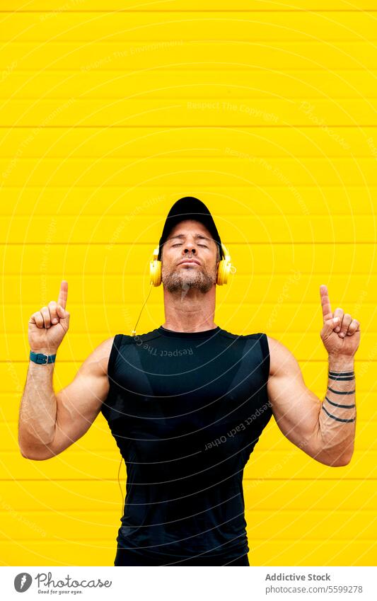 Ruhiger Sportler mit Kopfhörern, der über eine Mauer meditiert Athlet Augen geschlossen zuhören Musik Zeigen nach oben gelb Wand Windstille Porträt aussruhen