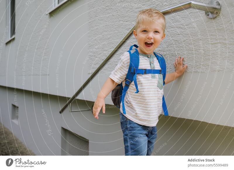 Happy niedlichen Kind Junge mit Rucksack stehen in der Nähe von grauen Wand Gebäude im Tageslicht Lächeln Reling heiter Kindheit spielerisch Weg Glück Sommer