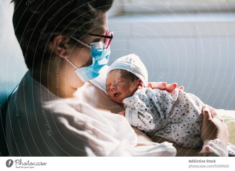 Eine Mutter trägt eine Gesichtsmaske, während sie ihr neugeborenes Baby im Krankenhauszimmer hält. Frauen Raum Anfänge Eltern Bonden anhänglich Anschluss Freude