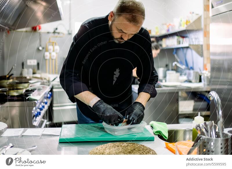 Bärtiger männlicher Profikoch mit schwarzen Handschuhen, der bei der Arbeit in einer Profiküche Lebensmittel in einen transparenten Behälter schichtet