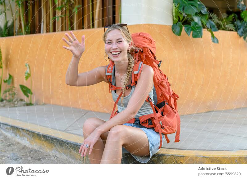 Fröhliche junge erwachsene Frau Tourist mit blonden langen Haaren orange Rucksack lässige Kleidung sitzen auf Stein Straße Grenze winkende Hand während des Wartens auf Freund und lachen gegen tropischen Hintergrund