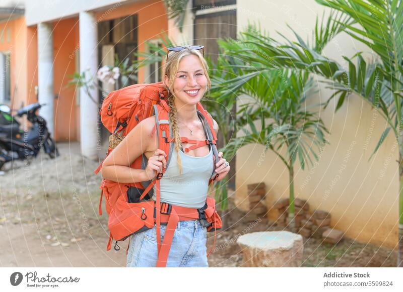 Glückliche junge blonde Frau steht mit Rucksack auf der Straße Abenteuer Backpacker schön verschwommener Hintergrund lässig heiter Textfreiraum tagsüber Freude