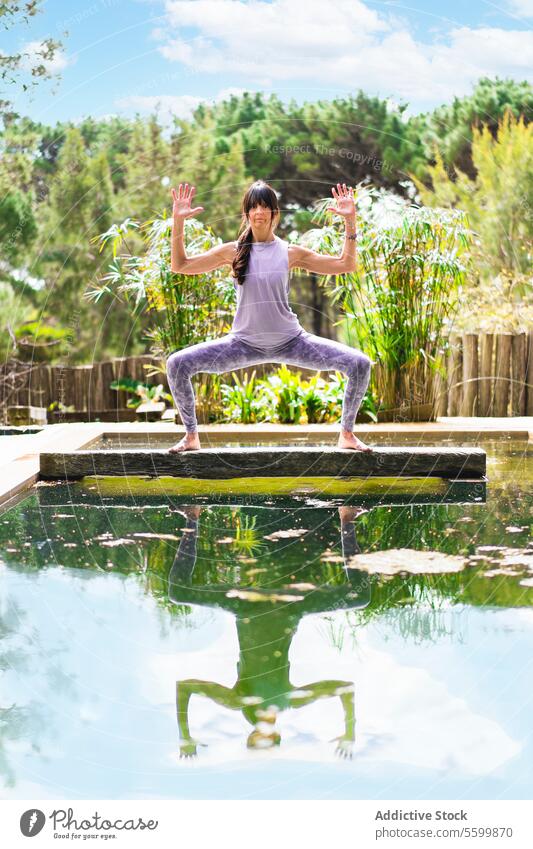 Eine Frau übt die Yogapose Göttin mit Kaktusarmen auf einem Baumstamm über einem Pool, Vertikalaufnahme Ästhetik Waffen Gleichgewicht Brücke Ruhe Konzentration