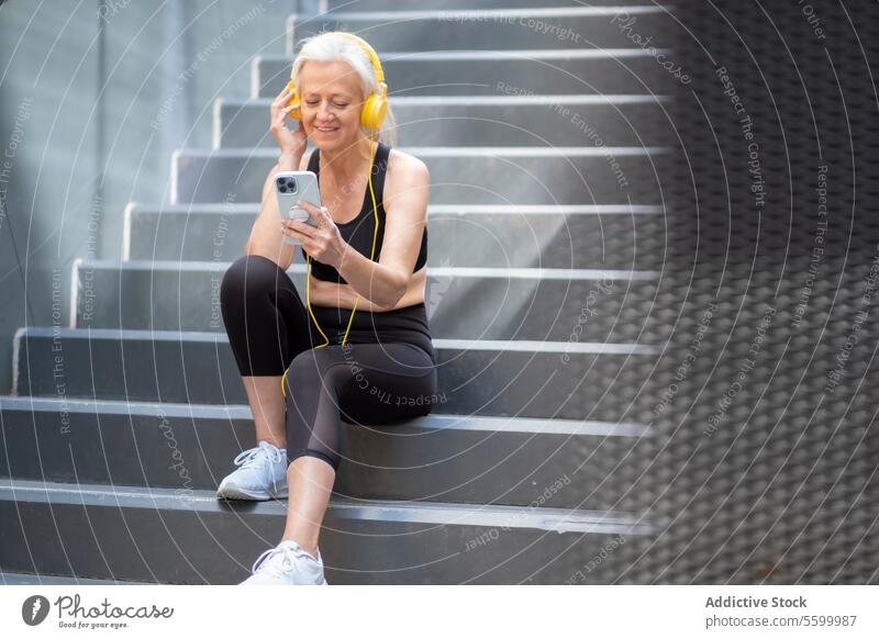 Ältere Frau genießt Musik auf Smartphone im Freien in Trainingskleidung Senior Kopfhörer Treppe Sitzen aktiv Lifestyle Gesundheit urban Großstadt Freizeit