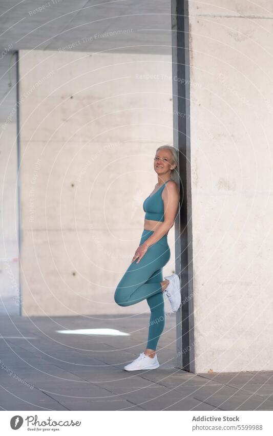Fitte ältere Frau in Sportkleidung lehnt an einer Wand Fitness Lächeln blaugrün fettarm Senior Pause urban Beton Übung Wellness Gesundheit reif sportlich