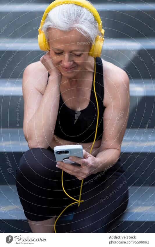 Ältere Frau genießt Musik über Kopfhörer im Freien in Trainingskleidung. Senior Smartphone Freitreppe Kleidung silbernes Haar Lächeln Freizeit Lifestyle