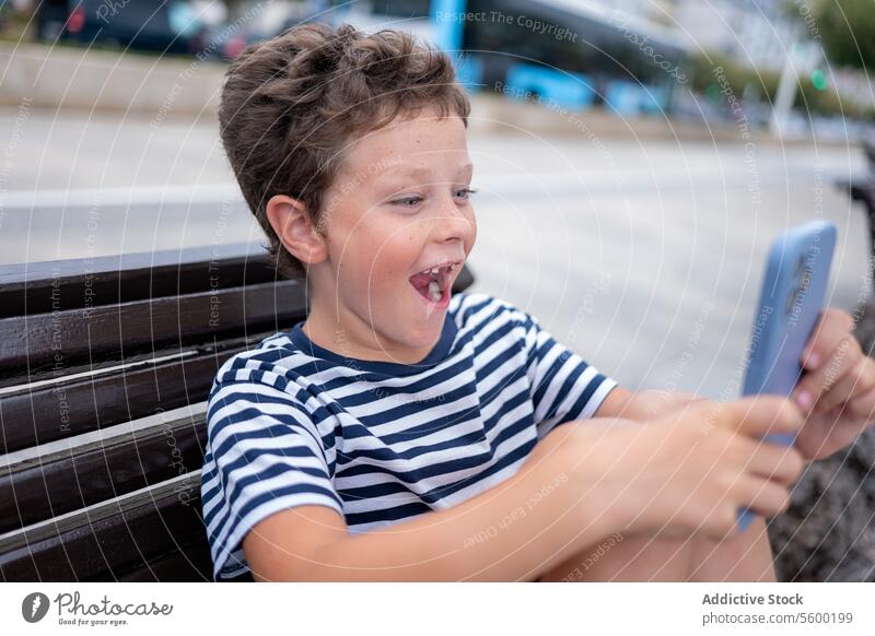 Seitenansicht eines jungen staunenden Jungen, der auf einer Bank am Meer sitzt und fröhlich ein Smartphone benutzt Seeküste Sitzen Glück erstaunt