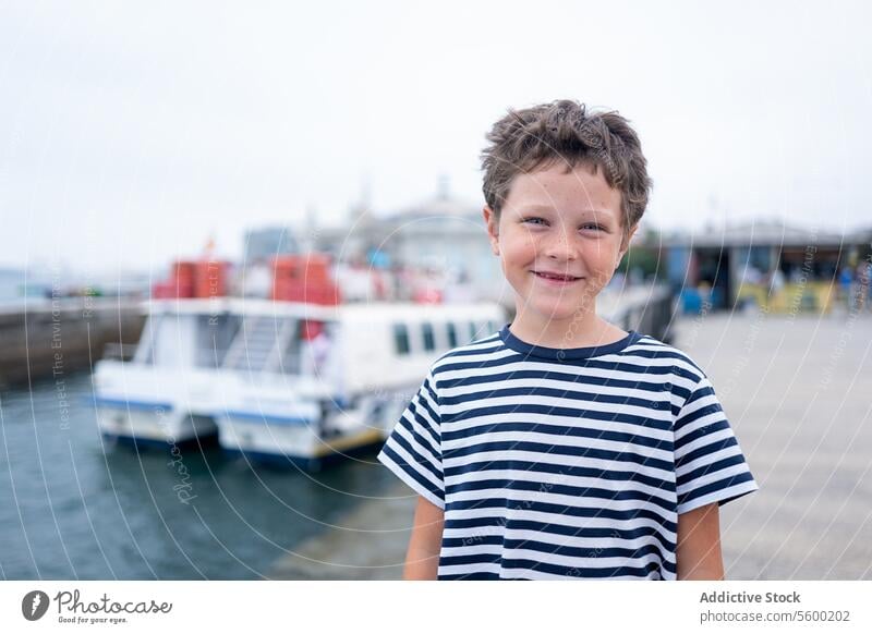 Fröhliches Kind im gestreiften T-Shirt lächelt in die Kamera mit dem Meer und Booten im Hintergrund während der Sommerferien Lächeln Glück MEER gestreiftes Hemd