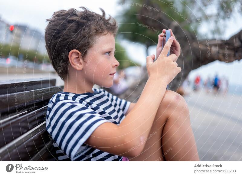 Seitenansicht eines Jungen, der auf einer Bank am Meer sitzt und fröhlich ein Smartphone benutzt Seeküste Sitzen Glück Technik & Technologie im Freien Kind