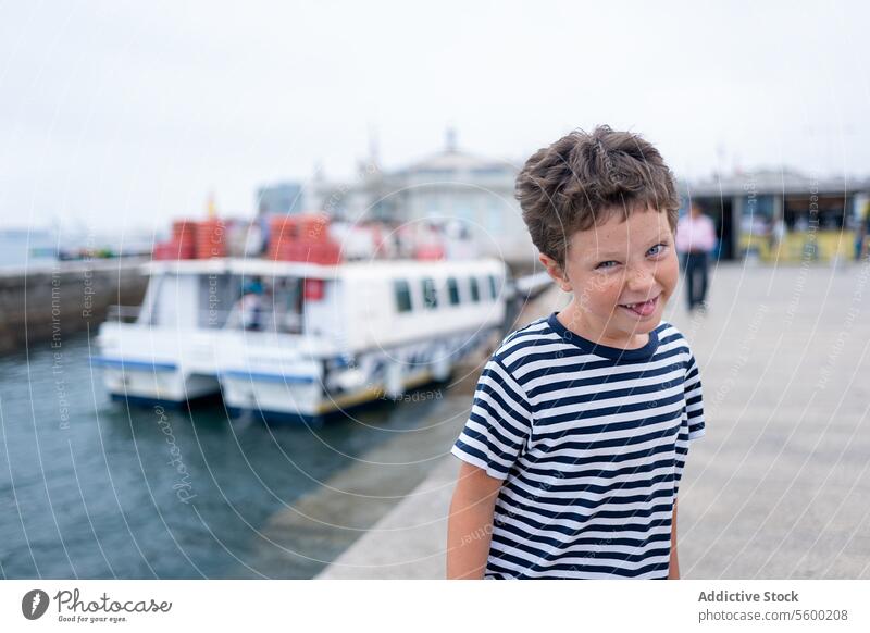 Fröhliches Kind im gestreiften T-Shirt lächelt in die Kamera, während es die Zunge herausstreckt, mit dem Meer und Booten im Hintergrund während der Sommerferien