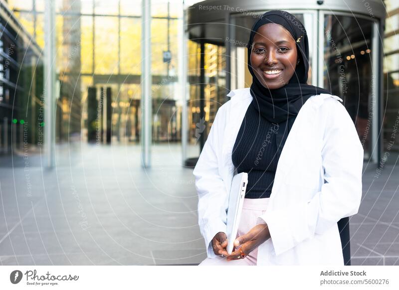 Fröhliche muslimische afroamerikanische Unternehmerin in Hijab und Formalien gekleidet, die einen Laptop hält und in die Kamera schaut, während sie auf der Straße steht