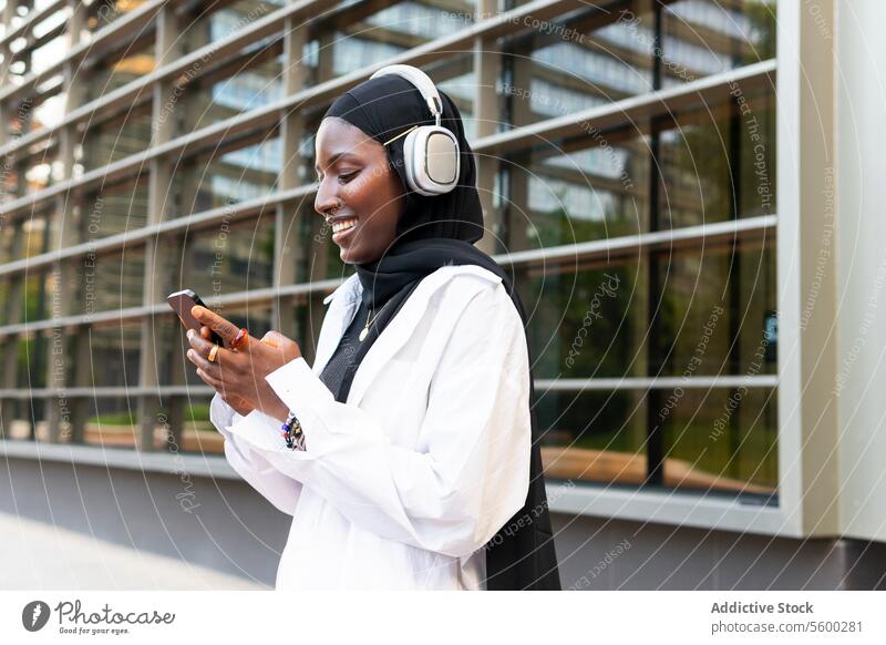 Glückliche Frau mit Kopfhörern und Mobiltelefon in der Stadt Geschäftsfrau Kopftuch zuhören Musik Smartphone heiter Hijab Lächeln muslimisch Großstadt