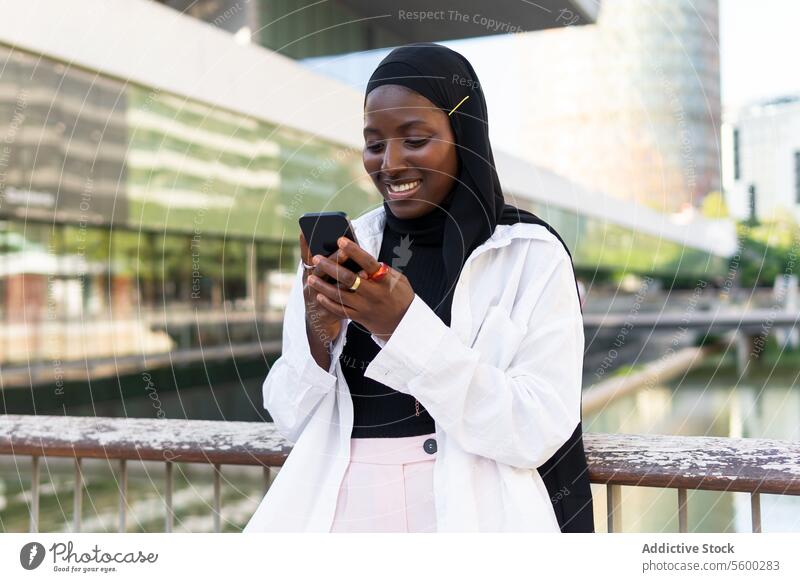 Fröhliche afroamerikanische junge Geschäftsfrau im Hidschab, die auf ihrem Smartphone surft, während sie vor einem modernen Gebäude steht Hijab Handy Frau
