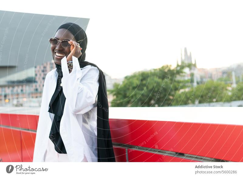 Glückliche Unternehmerin im Hidschab mit Brille in der Stadt Geschäftsfrau Hijab Sonnenbrille stylisch heiter muslimisch Tradition schwarz Kopftuch Schutzbrille