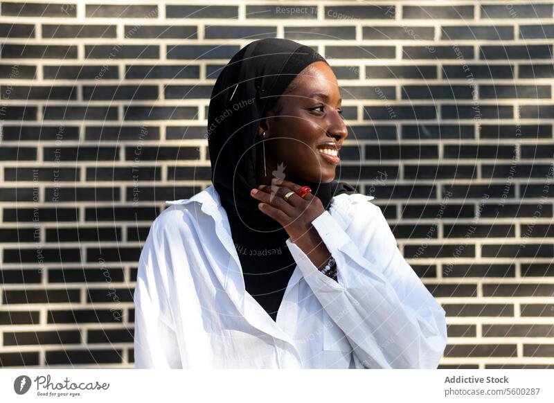 Porträt einer zuversichtlich lächelnden afroamerikanischen muslimischen Unternehmerin in schwarzem Hidschab und weißer Formalität, die an einer Backsteinmauer in der Stadt steht