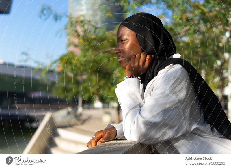 Seitenansicht einer nachdenklichen afroamerikanischen muslimischen Geschäftsfrau in schwarzem Hidschab, die ihr Kinn berührt und wegschaut, während sie sich auf ein Geländer in der Stadt stützt