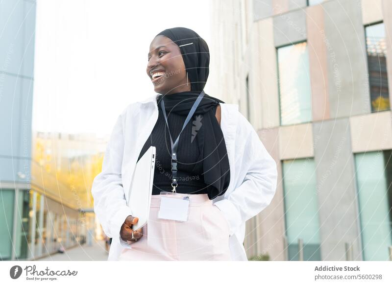 Fröhliche muslimische afroamerikanische Unternehmerin in Hijab und Formalien gekleidet, die einen Laptop hält und wegschaut, während sie auf der Straße steht