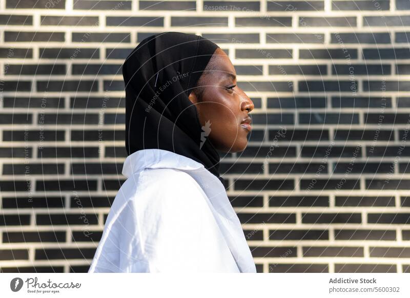 Porträtseitenansicht einer selbstbewussten afroamerikanischen muslimischen Unternehmerin in schwarzem Hidschab und weißer Formalität, die an einer Mauer in der Stadt steht