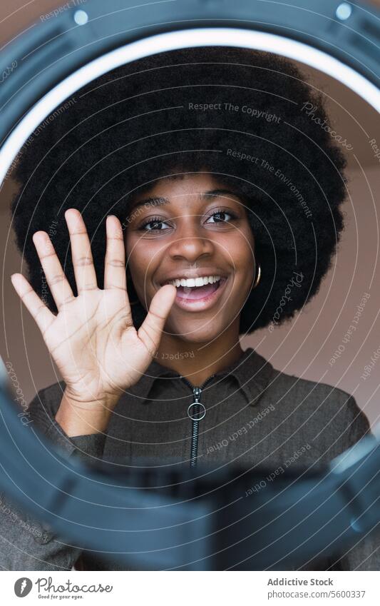 Zufriedene schwarze Frau winkt mit der Hand durch eine Ringleuchte vlog Blogger Ringlampe Wellenhand Gruß soziale Netzwerke Afro-Look Beleuchtung Lächeln Frisur
