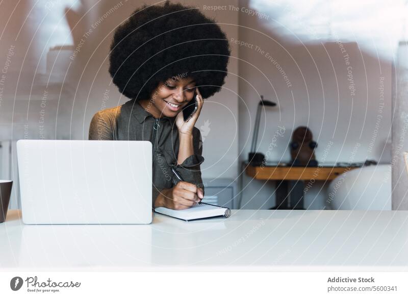 Glückliche schwarze Frau schreibt in ein Notizbuch neben einem Laptop Arbeit freiberuflich Arbeitsplatz Telefonanruf Notebook zur Kenntnis nehmen sprechen