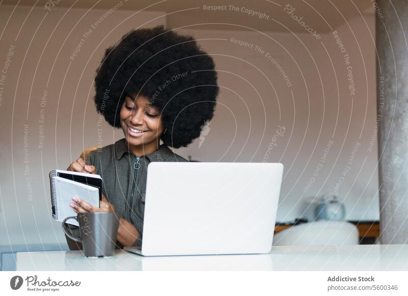 Fröhliche schwarze Frau schreibt in ein Notizbuch neben einem Laptop Arbeit freiberuflich Arbeitsplatz Notebook zur Kenntnis nehmen online Agenda Zeitplan